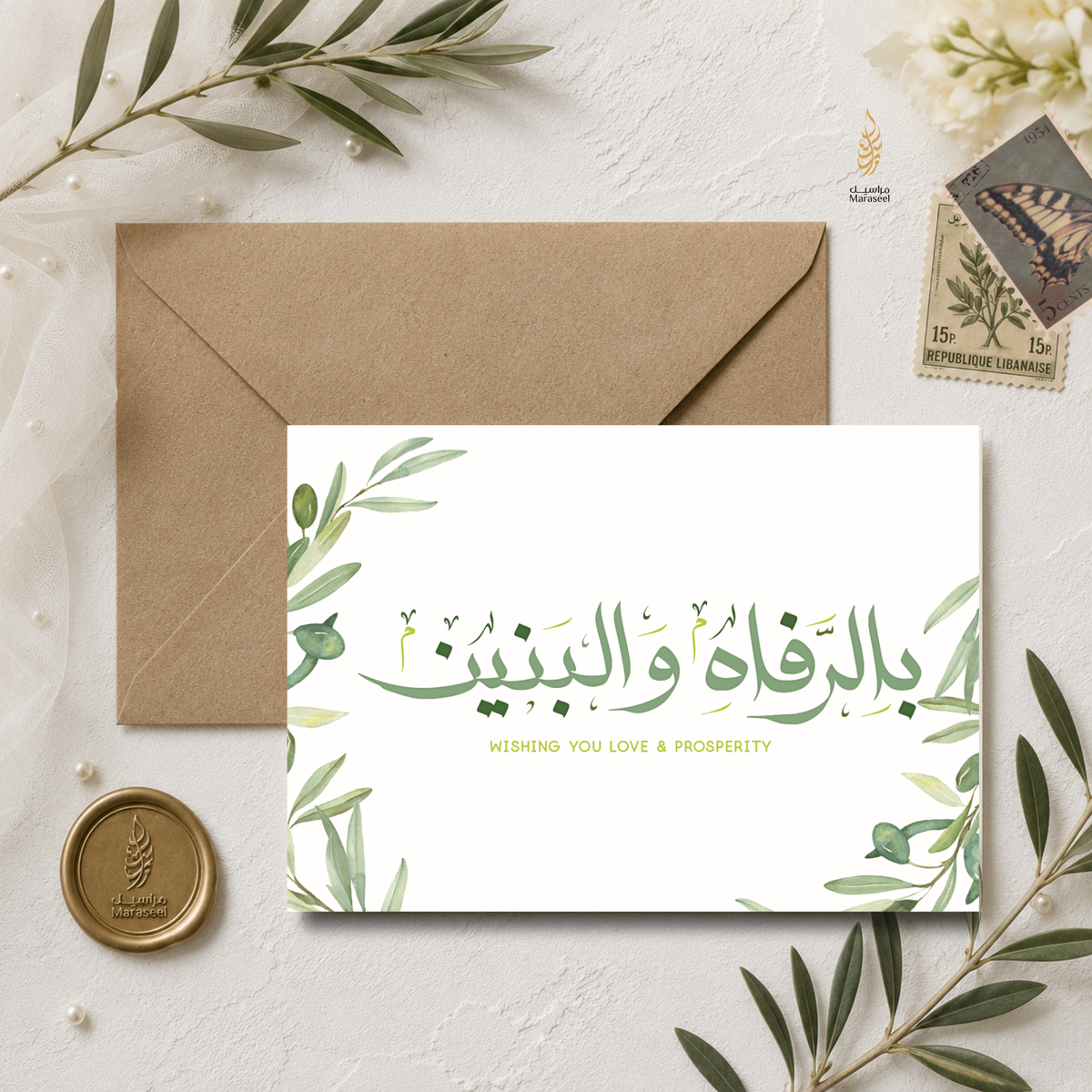 A white greeting card with green botanical illustrations and Arabic text, placed in front of a brown kraft envelope on a light textured surface. Decorative olive branches, a gold wax seal, and a vintage-style postage stamp surround the card.