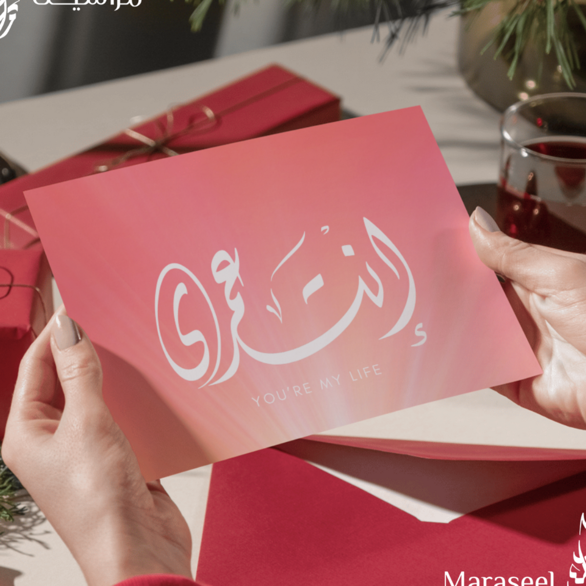 Person holding a red card with Arabic text and 'Maraseel' branding, surrounded by festive decorations.