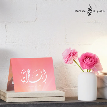 Decorative card with Arabic calligraphy on a pink background next to a vase with pink flowers.