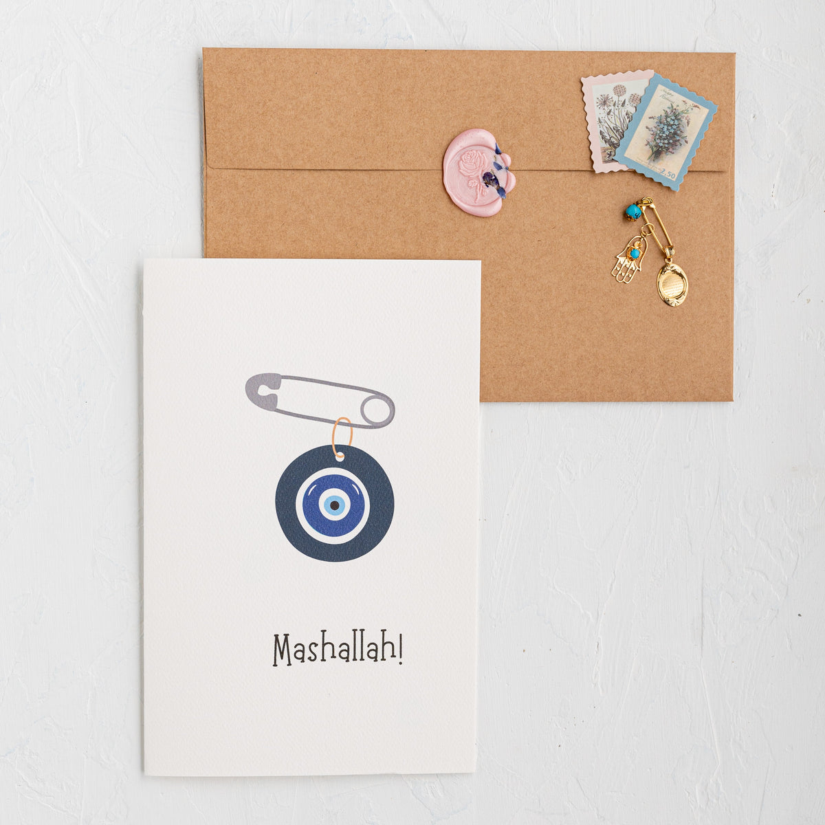 Card with a safety pin and target design, labeled 'Mashallah', on a white surface with a brown envelope.