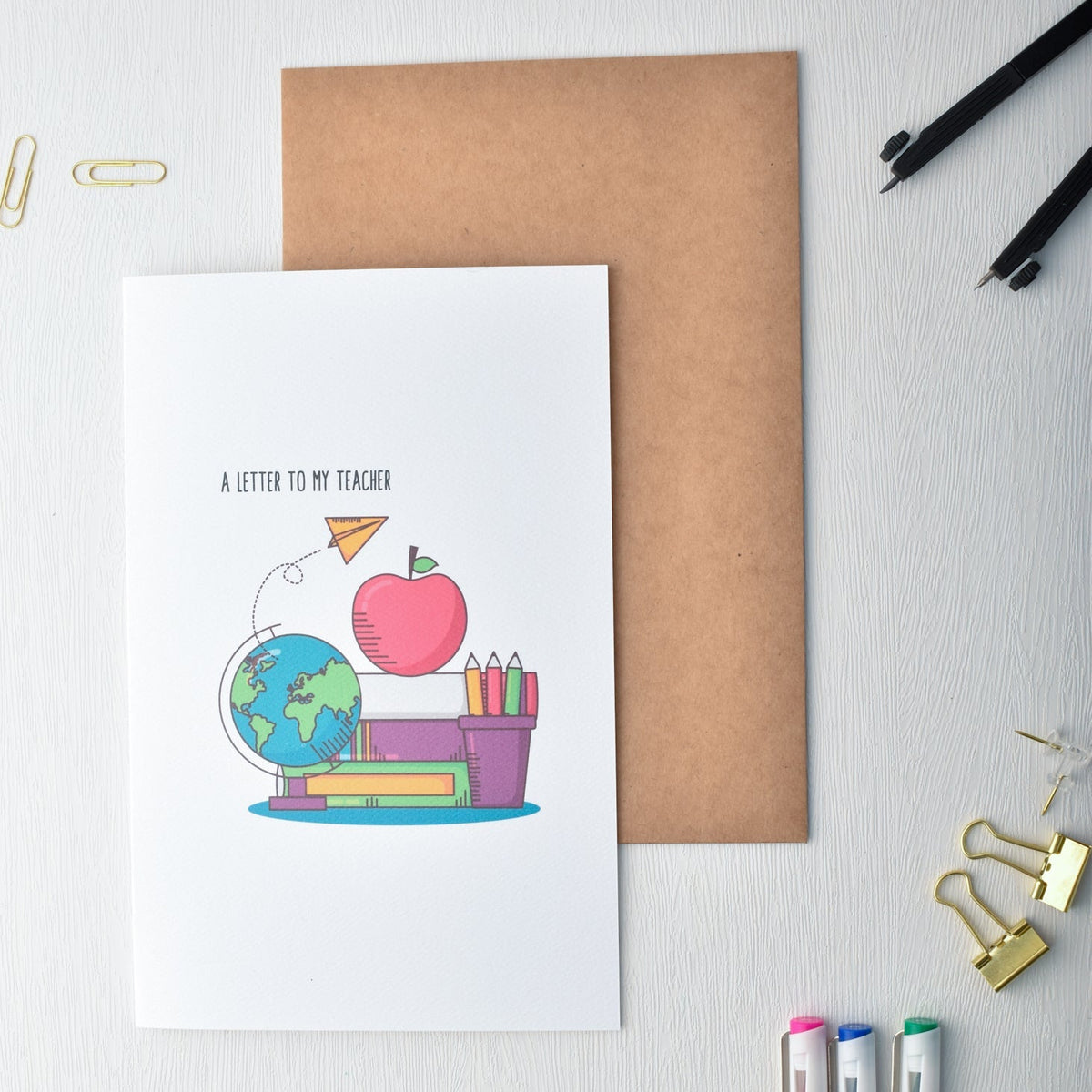Greeting card with illustration of a cartoon character, apple, globe, and books on a white surface with stationery items.