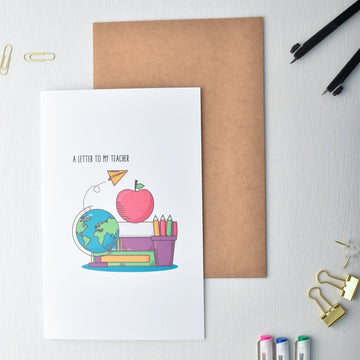 Greeting card with illustration of a cartoon character, apple, globe, and books on a white surface with stationery items.