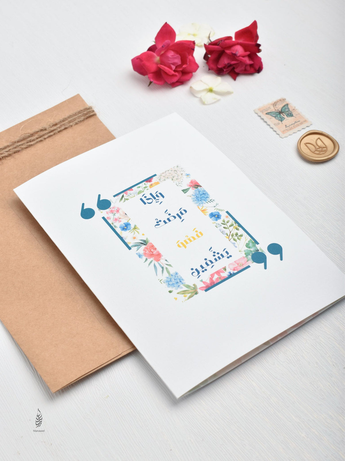 Greeting card with floral design and text, placed on a white surface with a brown envelope and red flowers.