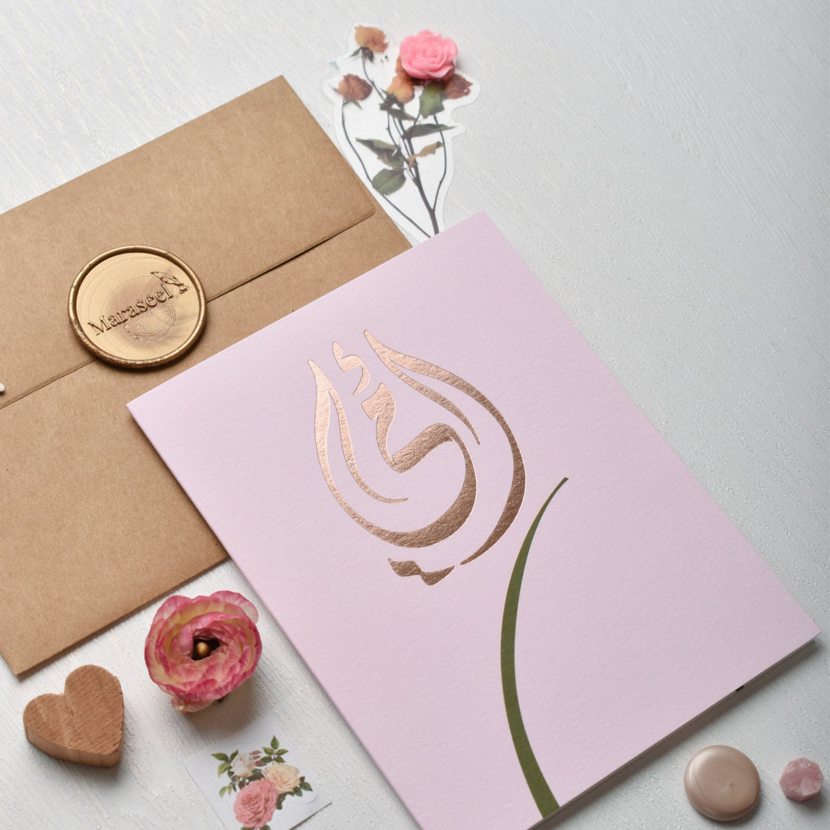Card with gold rose design on pink background, accompanied by a brown envelope and decorative items | Maraseel 