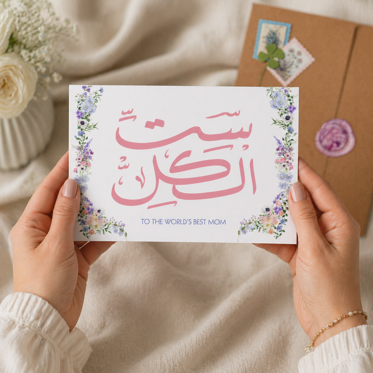 Hands holding a card with Arabic text and floral design, placed on a soft fabric background.