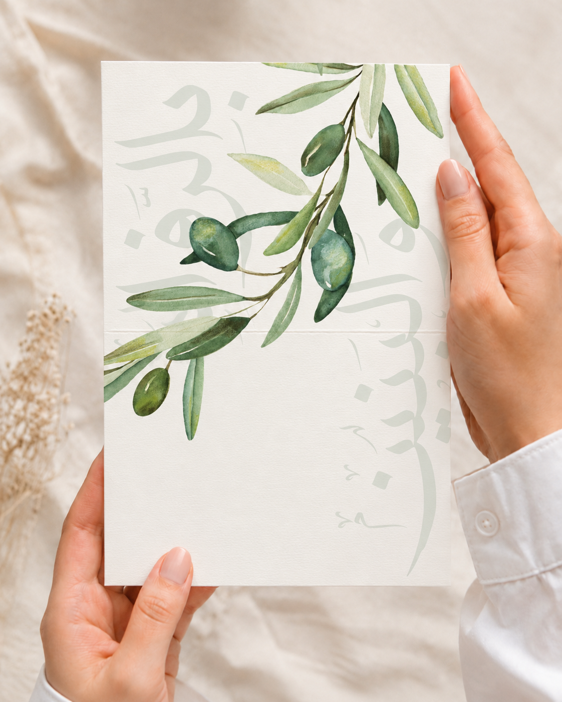 A person holds a square white art print featuring a watercolor illustration of green olive branches and leaves, with faint Arabic calligraphy in the background. The hands and white shirt cuff are visible against a soft beige fabric backdrop.