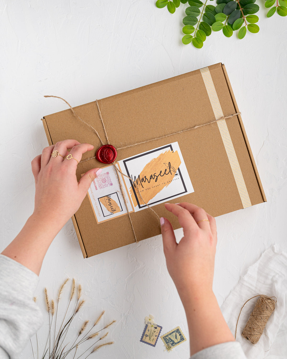 A person is writing on a card with a wax seal stamp, which is placed on a brown cardboard box with a window showing the card inside.